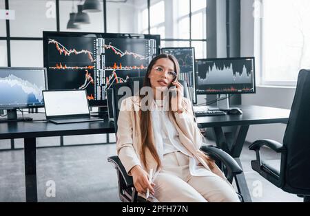 Talks by phone. Young female stock brocker works indoors in the office. Many of displays Stock Photo