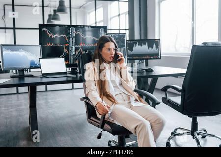 Talks by phone. Young female stock brocker works indoors in the office. Many of displays Stock Photo