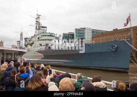 Tourist boat complete with tourists viewing HMS Belfast which is a Town-class light cruiser, built for the Royal Navy. Now permanently moored as a museum ship on the River Thames in London, UK. (133) Stock Photo