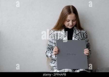 A 10-year-old girl is sitting, holding a gray leather photo book with an imprint of a memory in her hands Stock Photo