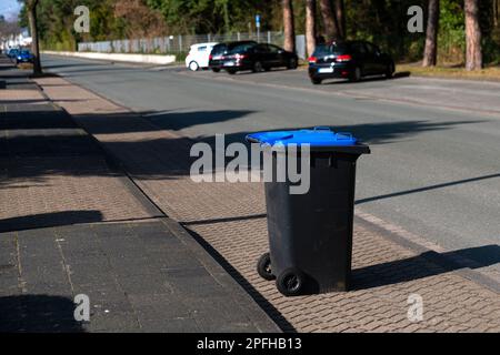 A black trash can with a blue lid on the edge of the street. Parked cars in the background. Stock Photo