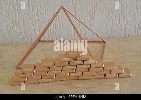 Chocolate. Handmade chocolate bars wrapped with golden paper in pyramid shape on a travertive marble table with copper pyramid in the background , Br Stock Photo