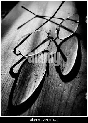 Black and white photo of a pair of eyeglasses with an elongated shadow on a rustic wooden table Stock Photo