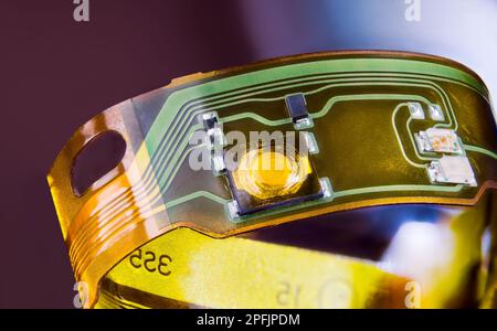 Electronic flexible printed circuit on plastic strip curled to ring on purple background. Green-yellow lines and small electric components on flex PCB. Stock Photo