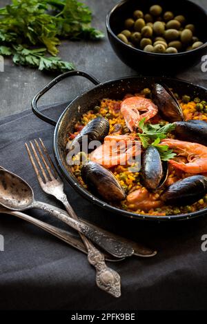 Paella in black pan with rice, shrimps, mussels, squid and meat, bowl with olives and vintage cutlery. Seafood paella, traditional spanish dish. Stock Photo