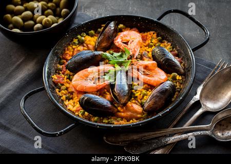 Paella in black pan with rice, shrimps, mussels, squid and meat, bowl with olives and vintage cutlery. Seafood paella, traditional spanish dish. Stock Photo