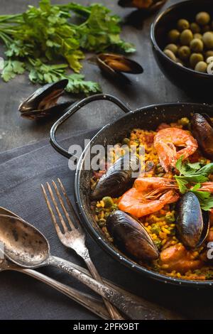 Paella in black pan with rice, shrimps, mussels, squid and meat, bowl with olives and vintage cutlery. Seafood paella, traditional spanish dish. Stock Photo