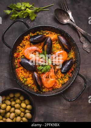 Paella in black pan with rice, shrimps, mussels, squid and meat, bowl with olives and vintage cutlery. Seafood paella, traditional spanish dish. Stock Photo