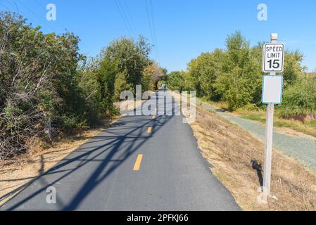 Bicycle and pedestrian path lined with trees on a sunny autumn day. A speed limit sign for cyclists is in foreground. Stock Photo