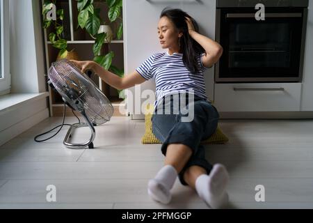 Pleased Asian woman blown by electric fan sits on floor enjoy cool wind from air ventilator in heat. Stock Photo