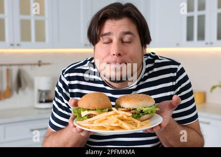 Happy overweight man holding plate with tasty burgers and French fries in kitchen Stock Photo