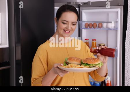 Happy overweight woman with ketchup and burgers near fridge in kitchen Stock Photo