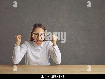 Stressed young woman with eyes closed screaming clenching fist sitting at desk Stock Photo