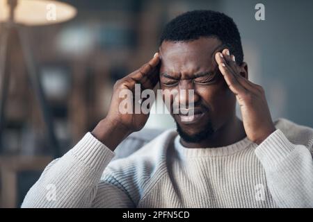This headache is unbearable. a handsome young man sitting alone in his living room and suffering from a headache. Stock Photo