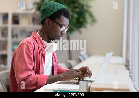 Young African American man freelancer working on laptop using public library as workspace Stock Photo