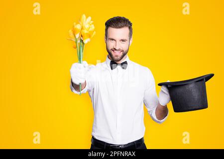 Portrait of romantic positive magician with bristle and modern hairstyle getting a yellow flowers from tophat and giving it front, isolated on grey Stock Photo