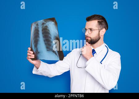 Portrait of pensive minded doc in white lab coat, uniform holding hand on chin looking at x-ray of lungs, trying to establish a diagnosis, isolated on Stock Photo