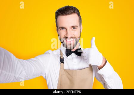 Self portrait of attractive cheerful man in white shirt with black bow, modern hairstyle, shooting selfie on front camera and gesture thumb up Stock Photo