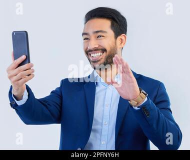 Smartphone, video call and asian businessman in studio with wave, talking and networking on white background. Phone, conversation and b2b Stock Photo