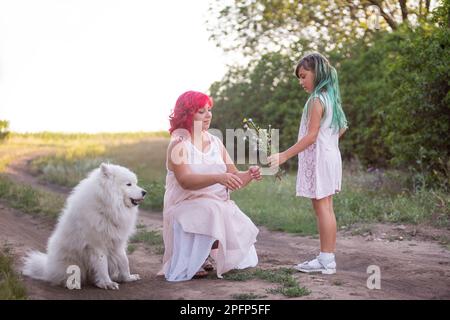 Girl with green hair gives wild flowers to mother with pink. Traveling with Samoyed dog, pets to nature. The tenderness of motherhood, one parent. Div Stock Photo