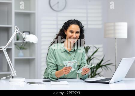 A young Muslim woman is sitting in the office at a desk. She holds cash in her hands, received a salary, a bonus, an award. She looks at the camera smiling. Stock Photo