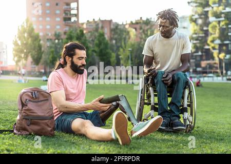 In a sunny park, two young adults with disabilities chat on the grass, one with a prosthetic leg and the other, an African man, in a wheelchair, shari Stock Photo