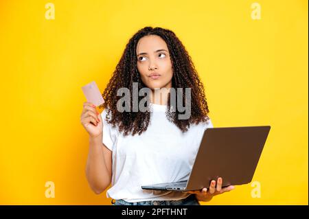 Pensive brazilian or hispanic woman with curly hair, wearing white t-shirt, holding laptop and bank credit card, looking away thoughtfully, standing over isolated yellow background Stock Photo
