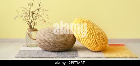 Vase with tree branches and knitted poufs on carpet near yellow wall in room Stock Photo