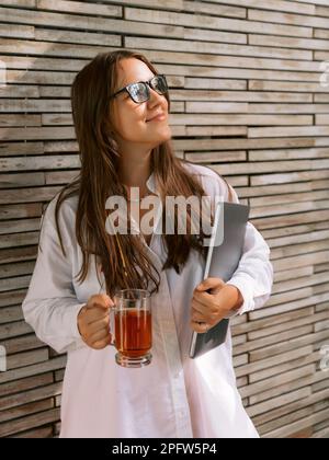 Young woman freelancer with glasses and holding laptop with cup of tea Stock Photo