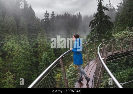 Canada Autumn travel destination in British Columbia. Asian tourist woman walking in famous attraction Capilano Suspension Bridge Park in North Stock Photo