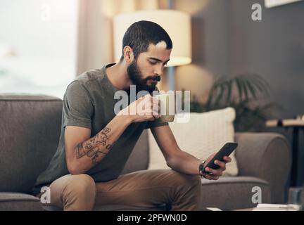Coffee breaks are for catching up. a young man having coffee and using a smartphone on the sofa at home. Stock Photo