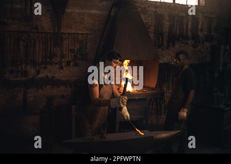 Strike while the iron is hot. a young woman hitting a hot metal rod with a hammer in a foundry. Stock Photo