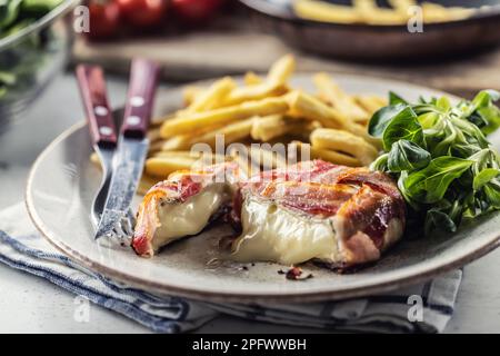 Cut melting cheese in a bacon wrap served on a plate with french fries and green salad. Stock Photo