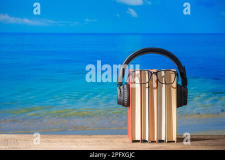 Set of books, headphones and eyeglasses on wooden table near sea water on the beach, close up. Audiobooks and education concept Stock Photo