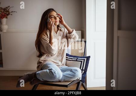 Serious pretty millennial mixed race woman calls by phone, talks, sits on chair, looks out window Stock Photo