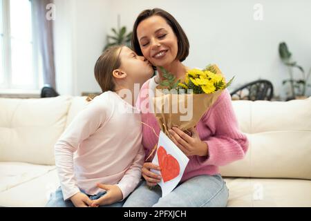 Cheerful little girl kissing european millennial woman with flowers hold postcard with heart in living room Stock Photo