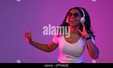 Happy Black Female Wearing Headphones And Stylish Sunglasses Posing In Neon Light Stock Photo
