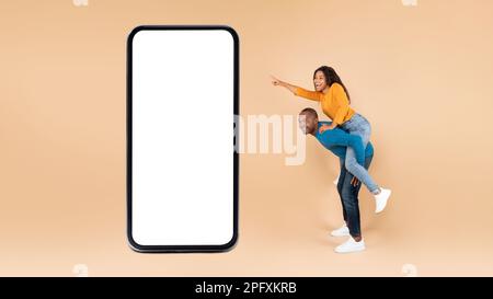 Great mobile app. Happy black couple presenting huge smartphone with mockup, man riding lady on back Stock Photo