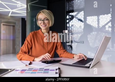 Portrait of successful financier woman, businesswoman behind paper accounting work smiling and looking at camera, female worker happy at workplace using laptop inside office, blonde in glasses. Stock Photo