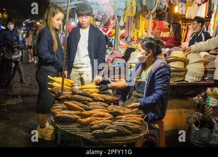 A young Vietnamese couple stop to buy grilled corn-on-the-cob at the bustling night market in Dalat, Vietnam. Stock Photo