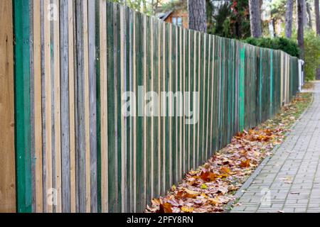 Fence of different colored wooden planks with autumn colored leaves and gray pedestrian pavement going to perspective view Stock Photo