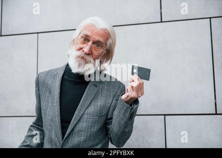 Senior businessman in formal clothes, with grey hair and beard is outdoors holds credit card Stock Photo