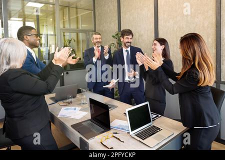Team of multiracial businesspeople wearing classy suits gathering at table with laptops in conference room and applauding after successful meeting Stock Photo