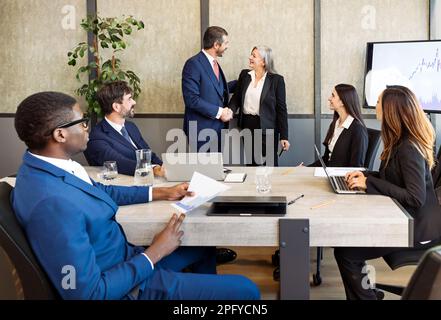 Group of diverse businesspeople in formal wear sitting at table and looking at coworkers shaking hands during business meeting in modern office Stock Photo
