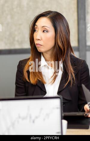Concentrated Chinese businesswoman in formal wear sitting at table and looking away thoughtfully during meeting Stock Photo