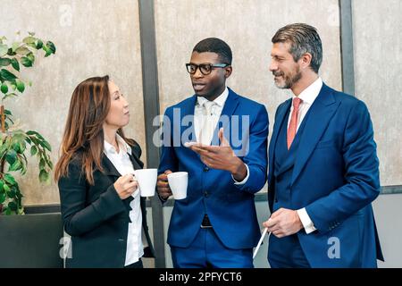 Multiethnic coworkers in classy clothes standing with cups of hot drinks and speaking while having break from work Stock Photo