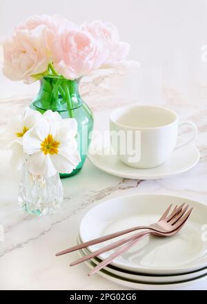 Delicate pink tulips with white primula flowers and dessert plates with rose gold forks on marble background.  Vertical format with focus on tulips. Stock Photo