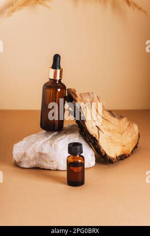 Brown cosmetic serum and essential oil bottles on a stone podium and wooden bark on neutral background. Natural skin care cosmetics concept. Closeup. Stock Photo