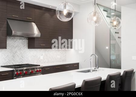 A modern kitchen detail with dark wood cabinets, stainless steel stove and hood, chairs sitting at a large island, and a herringbone backsplash. Stock Photo