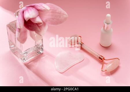 Rose quartz crystal facial roller and gua sha scraper, face serum and magnolia flower in vase on pink background. Facial massage kit for lifting thera Stock Photo